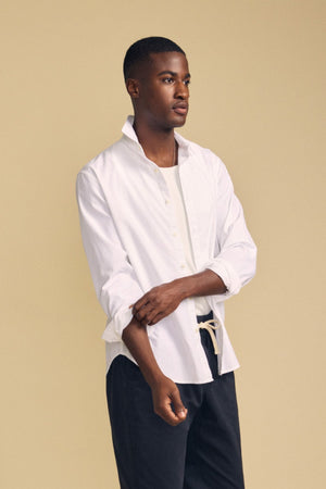 A man stands against a beige background, wearing the RICK SHIRT by Velvet by Graham & Spencer—a crisp white cotton button-up styled over a white top and dark pants, with one sleeve partially rolled up.