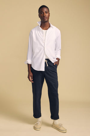 A person stands against a beige background wearing the Velvet by Graham & Spencer RICK SHIRT layered over a white undershirt, paired with dark drawstring pants and beige sneakers.