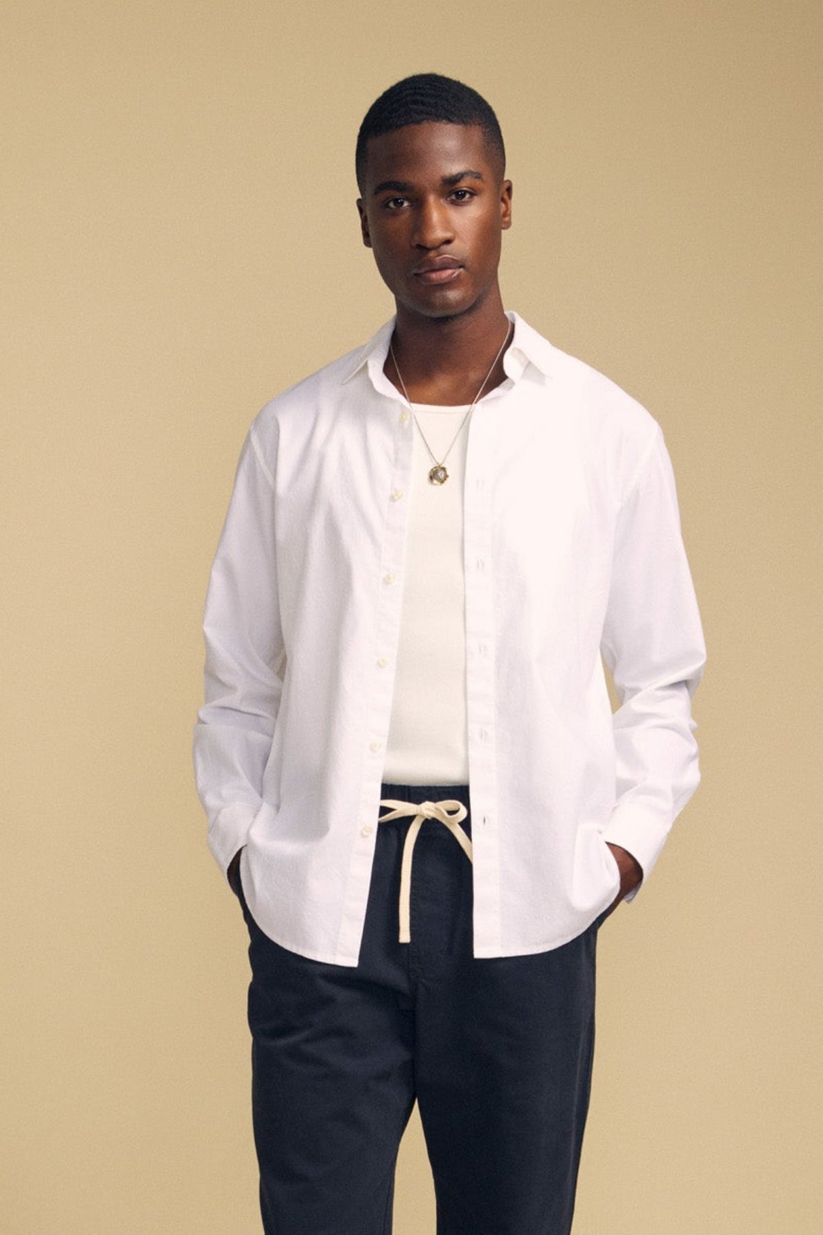A person stands against a beige background, wearing the crisp white RICK SHIRT by Velvet by Graham & Spencer layered over a white tank top, paired with dark pants, a necklace, and a rope-style belt, with hands in pockets.-42092506480833