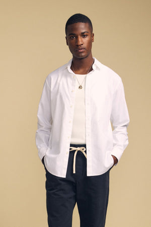 A person stands against a beige background, wearing the crisp white RICK SHIRT by Velvet by Graham & Spencer layered over a white tank top, paired with dark pants, a necklace, and a rope-style belt, with hands in pockets.