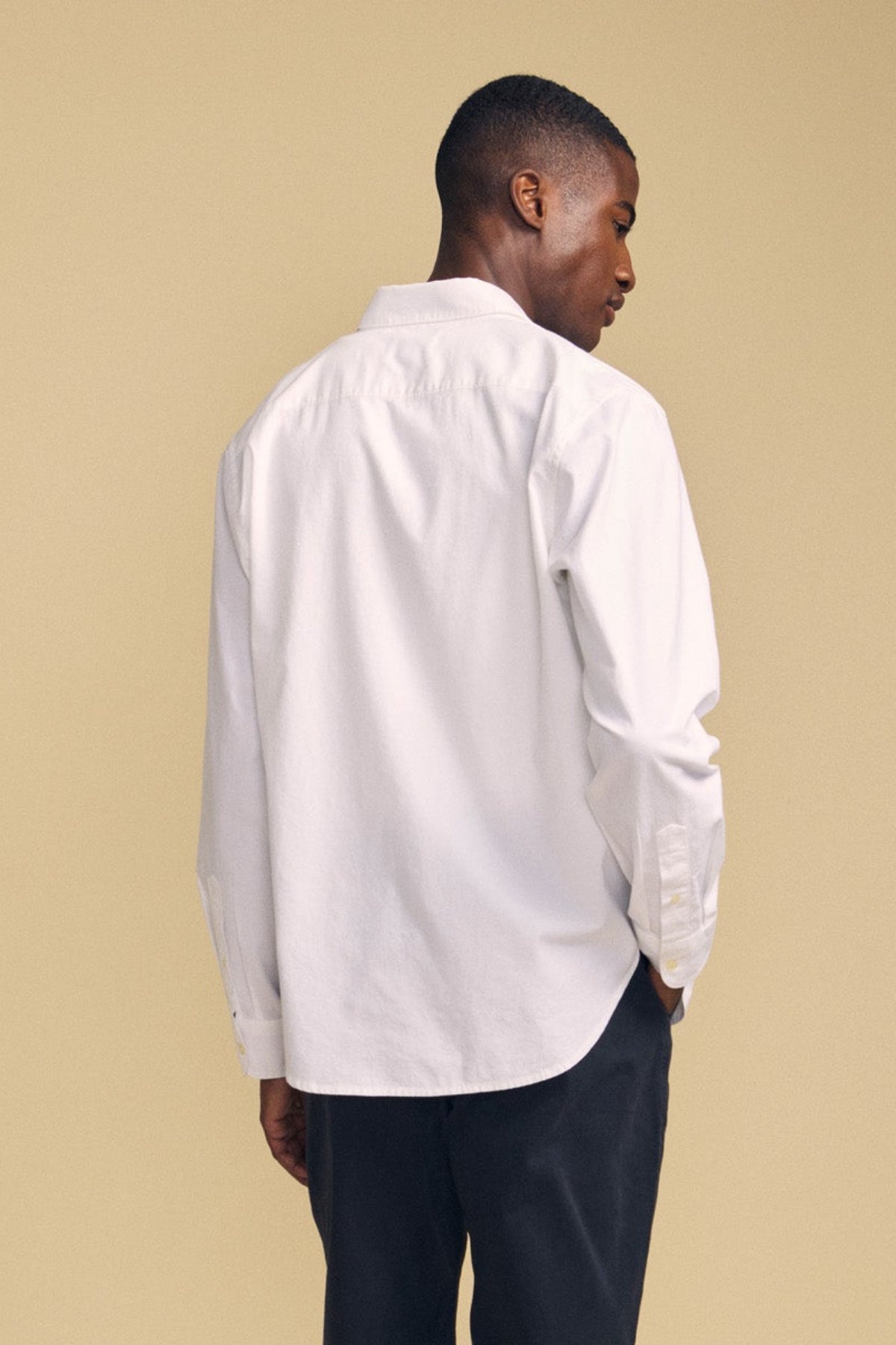 A person wearing the RICK SHIRT—a long-sleeve white cotton button-up by Velvet by Graham & Spencer—and dark pants stands with their back to the camera against a beige background.