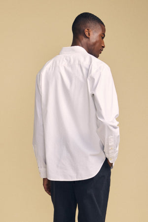 A person wearing the RICK SHIRT—a long-sleeve white cotton button-up by Velvet by Graham & Spencer—and dark pants stands with their back to the camera against a beige background.