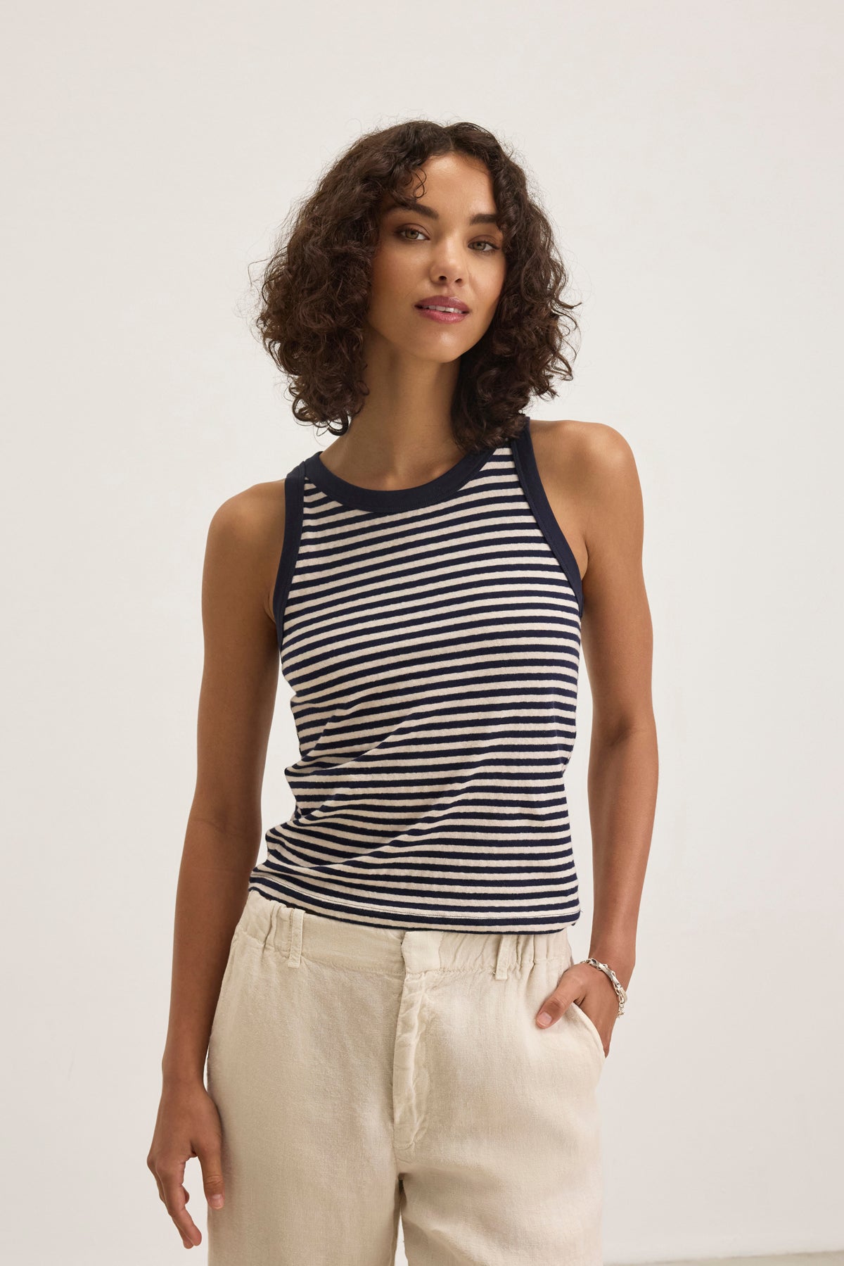   A person with curly hair wears the LUA TANK TOP by Velvet by Graham & Spencer, featuring black and white stripes, paired with beige pants against a plain background. 