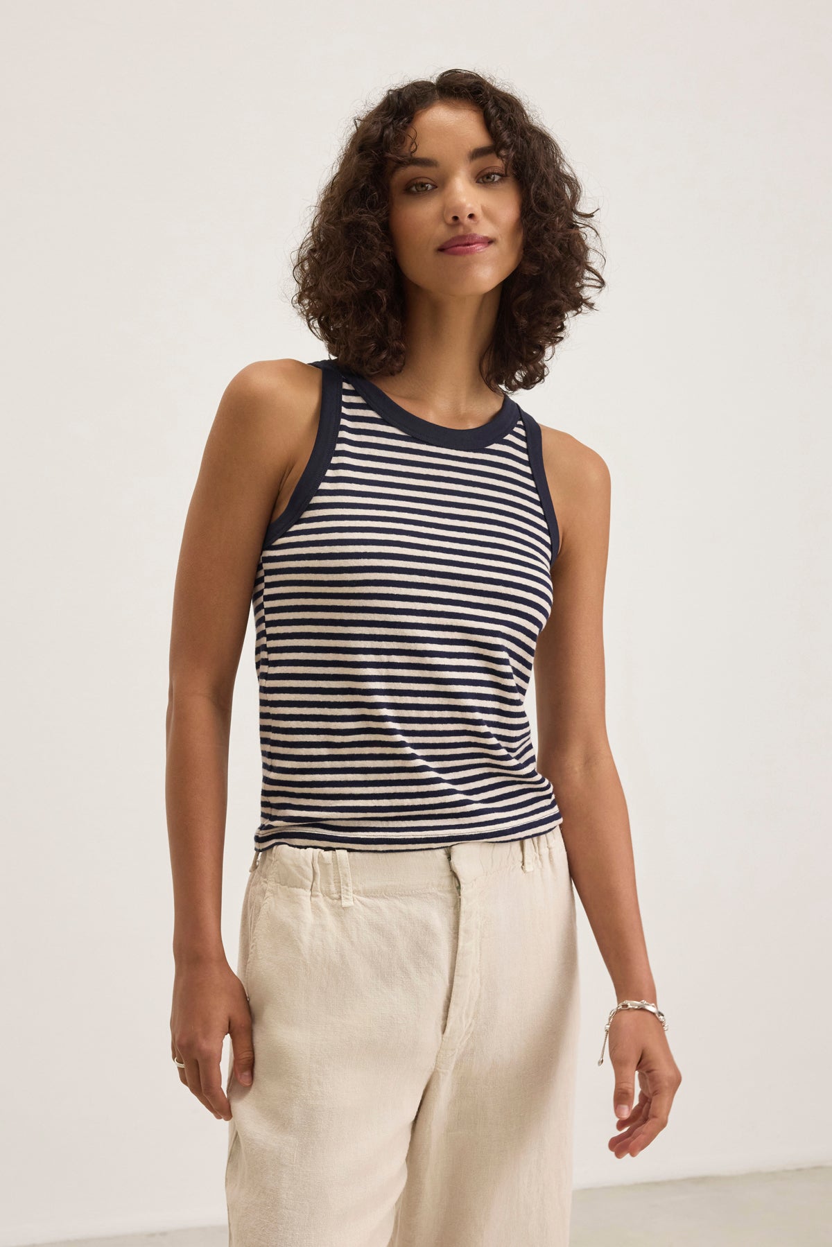   A person with curly hair wears a LUA TANK TOP by Velvet by Graham & Spencer, featuring black and white stripes, paired with beige pants against a plain background. 