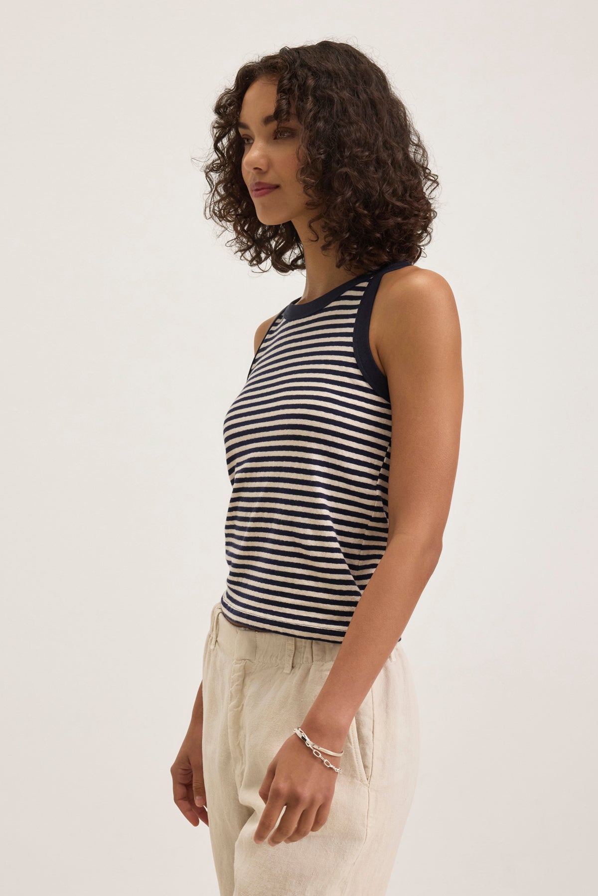   A person with curly hair wears the LUA TANK TOP, a black and white striped sleeveless piece by Velvet by Graham & Spencer, paired with beige pants against a plain background. 