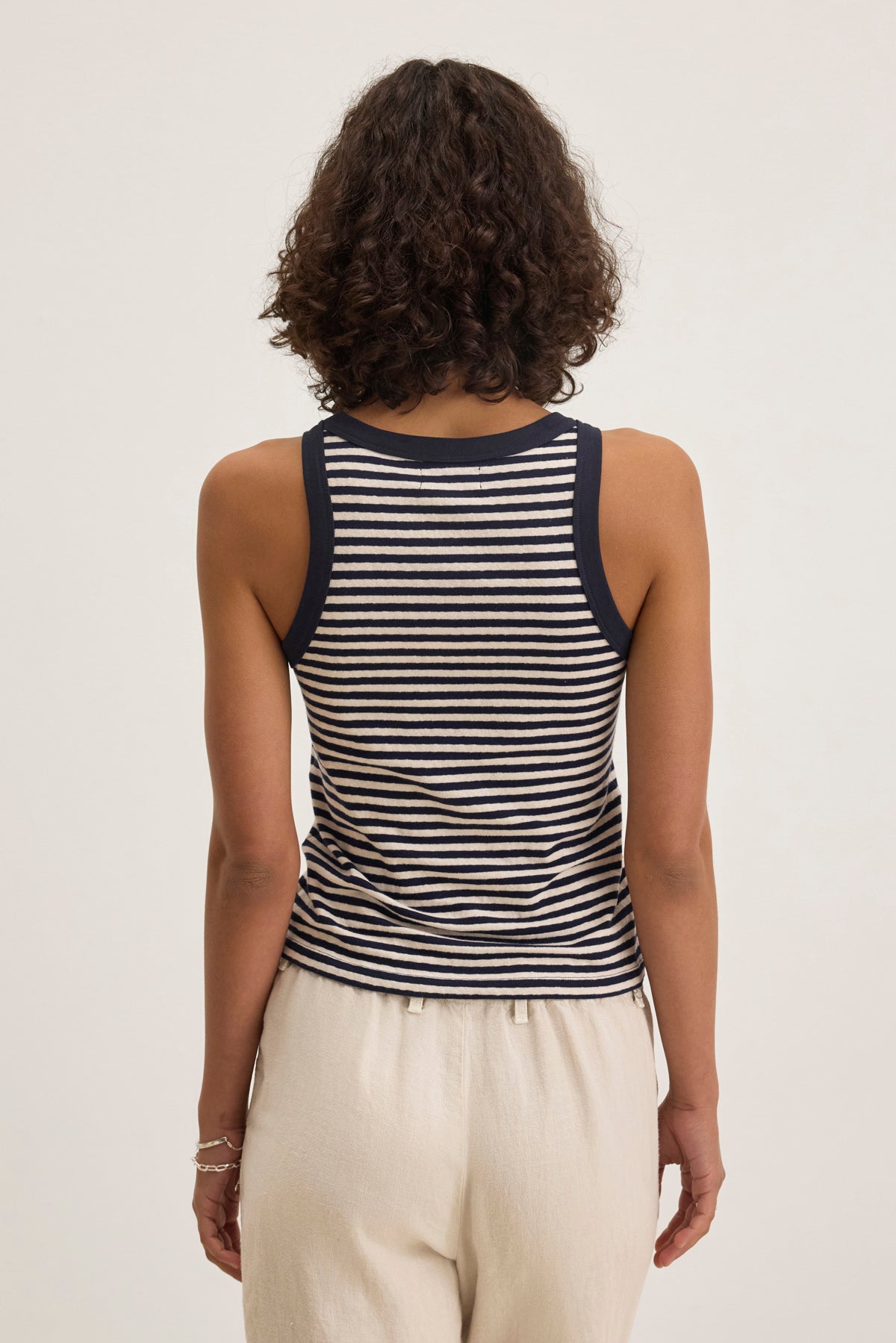   A curly-haired individual is wearing the LUA TANK TOP by Velvet by Graham & Spencer, featuring black-and-white stripes, paired with light-colored pants, and facing away. 