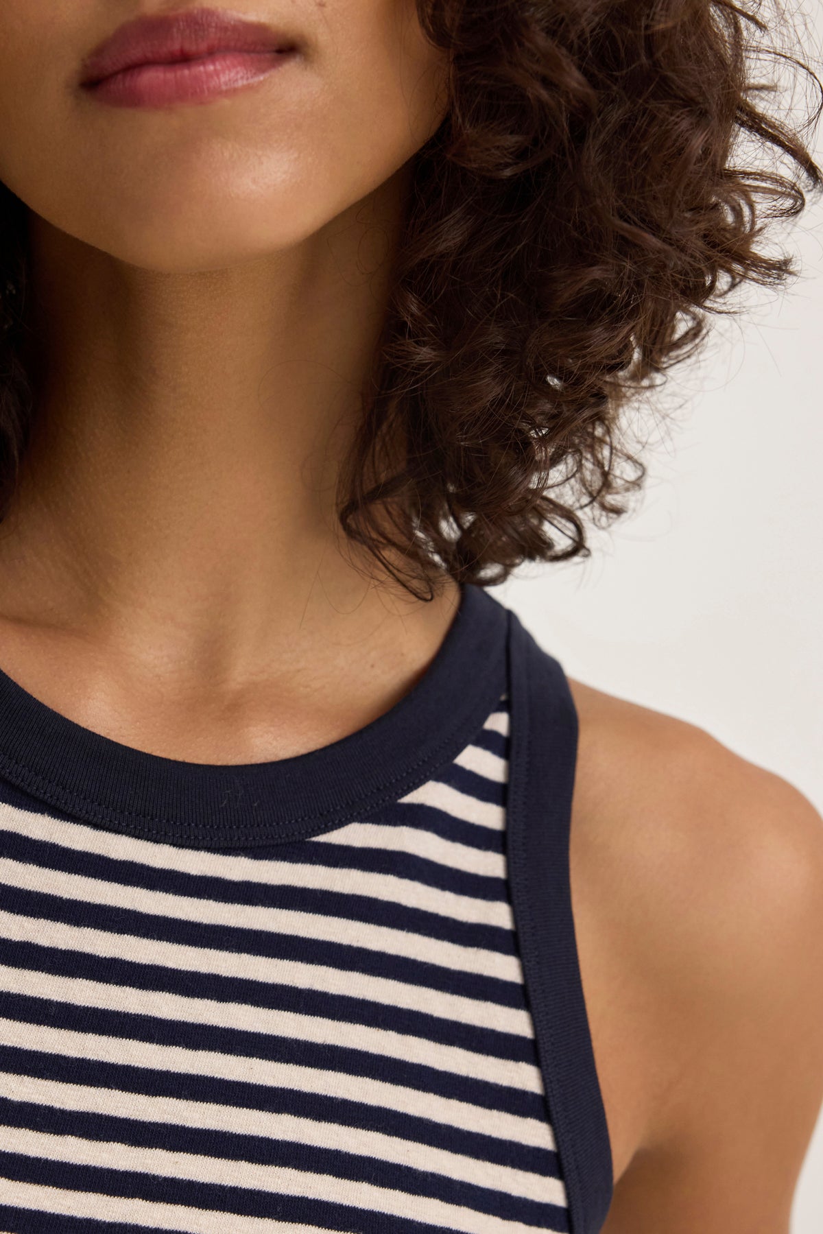   Someone with curly hair is wearing the LUA TANK TOP, a sleeveless navy and white striped top from Velvet by Graham & Spencer. 