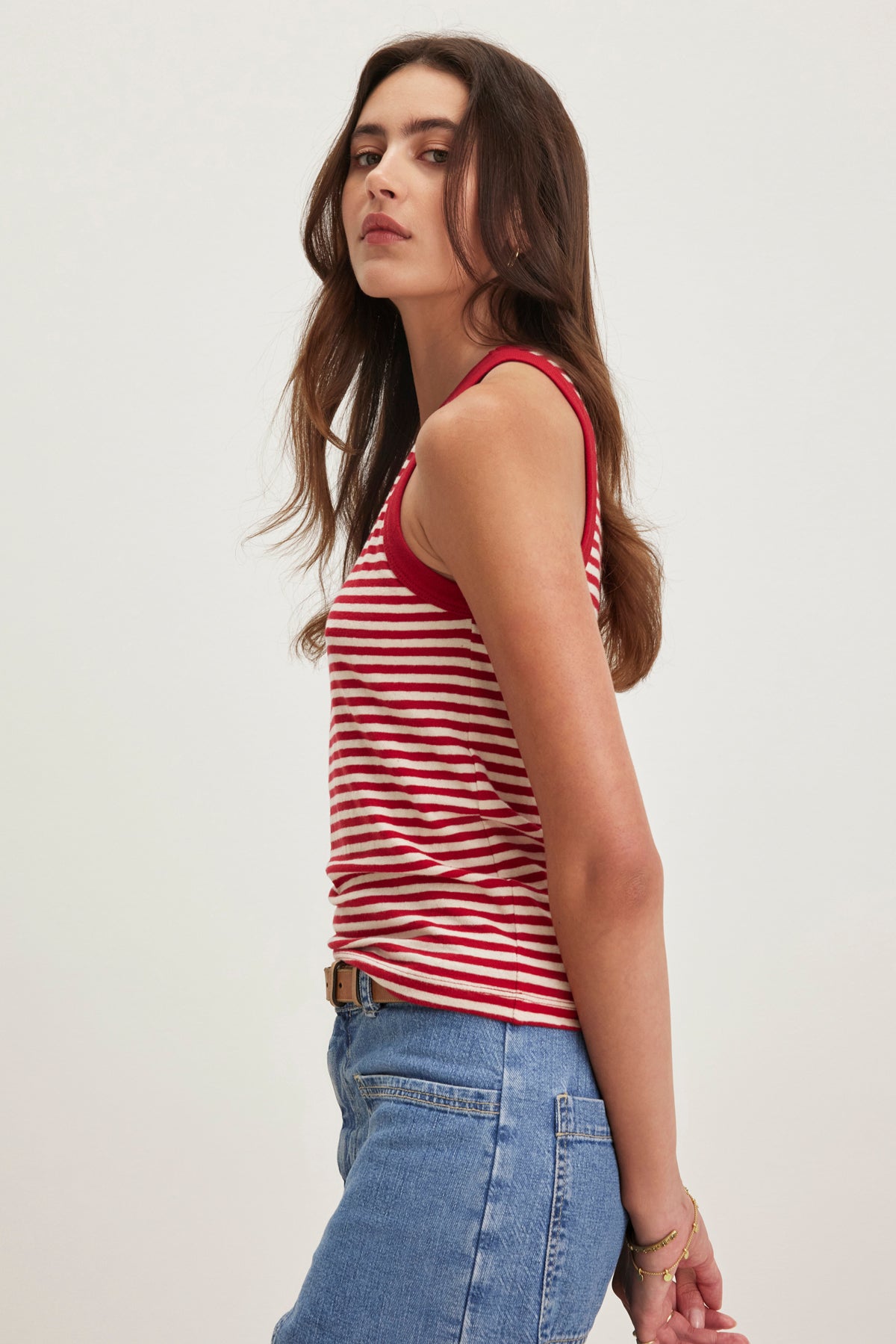   A person with long hair is wearing the LUA TANK TOP from Velvet by Graham & Spencer, which is red and white striped, sleeveless, and paired with blue jeans. They are looking to the side. 