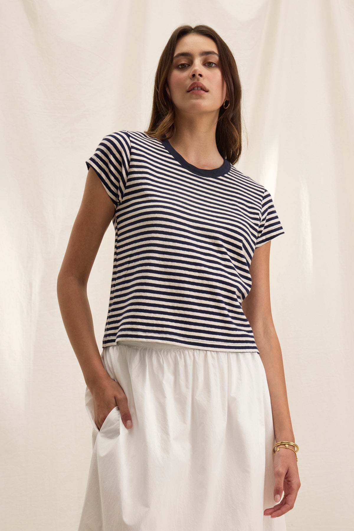 A person in a TASH TEE by Velvet by Graham & Spencer, featuring black and white stripes and a crew neckline, pairs it with a white skirt against a light background. Their right hand rests in a pocket, highlighting the casual elegance of cotton jersey fabric.-38820850335937