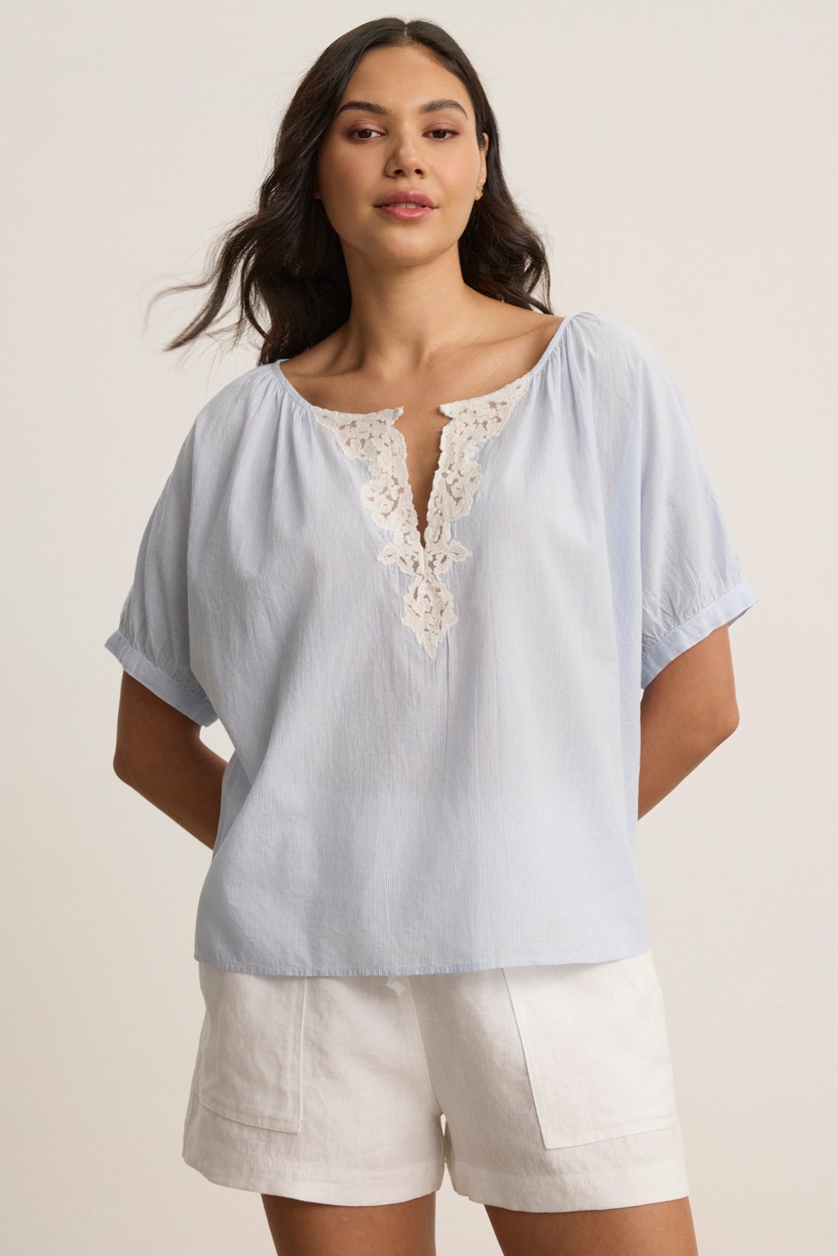 A woman wears the PENELOPE BLOUSE by Velvet by Graham & Spencer, a light blue boatneck top with lace detailing, paired with white shorts, standing against a plain background.