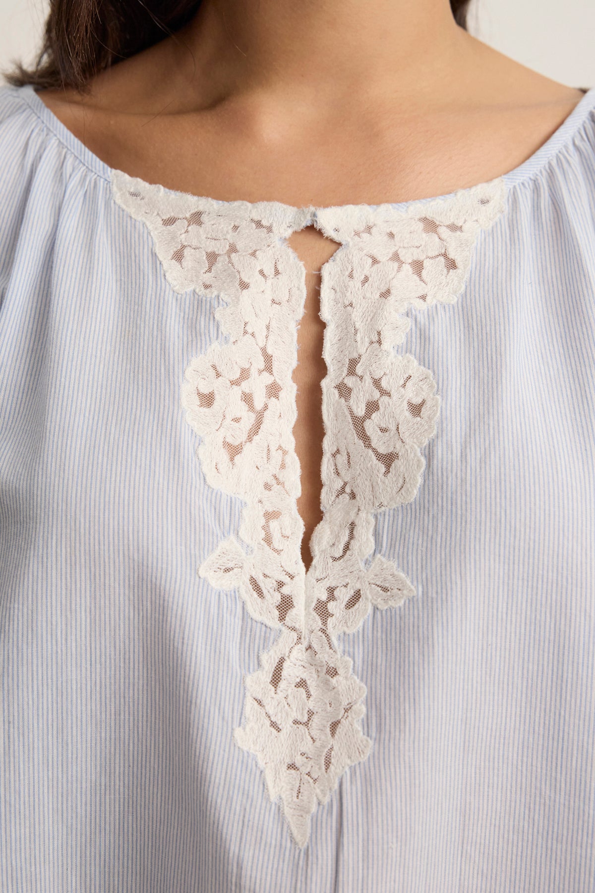 Close-up of a woman's upper torso in the PENELOPE BLOUSE by Velvet by Graham & Spencer—a light blue, yarn-dyed blouse with pinstripes and a V-shaped neckline featuring white lace accents.-Hover Image