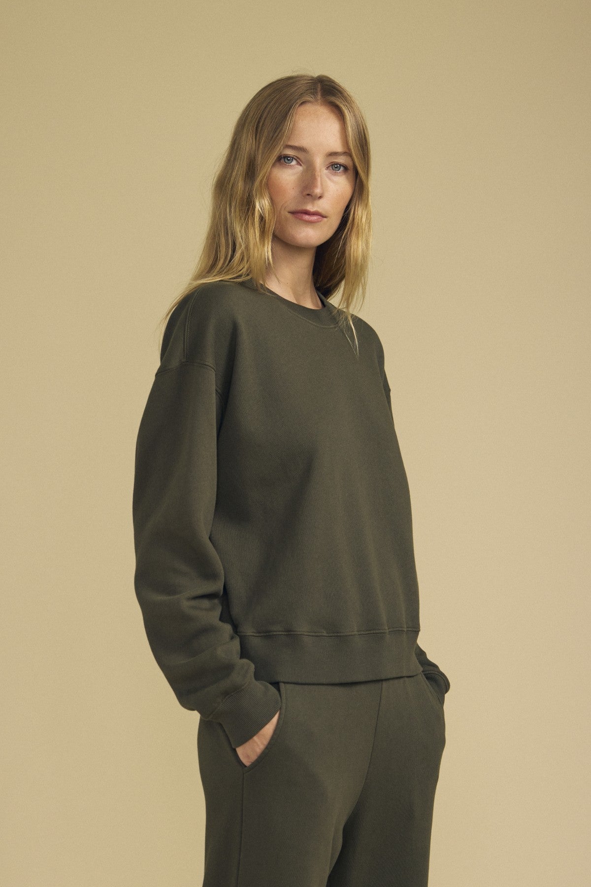 A woman with long blonde hair stands with her hands in her pockets against a plain beige background, showcasing a relaxed silhouette while wearing an olive green cotton fleece YOKO Sweatshirt and matching pants by Velvet by Graham & Spencer.-41838118404289