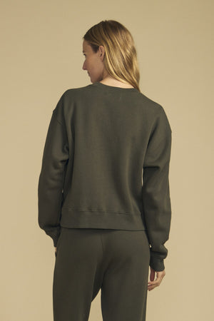 A person stands with their back to the camera, wearing a dark green Velvet by Graham & Spencer YOKO Sweatshirt and matching pants in a relaxed silhouette against a plain beige background.