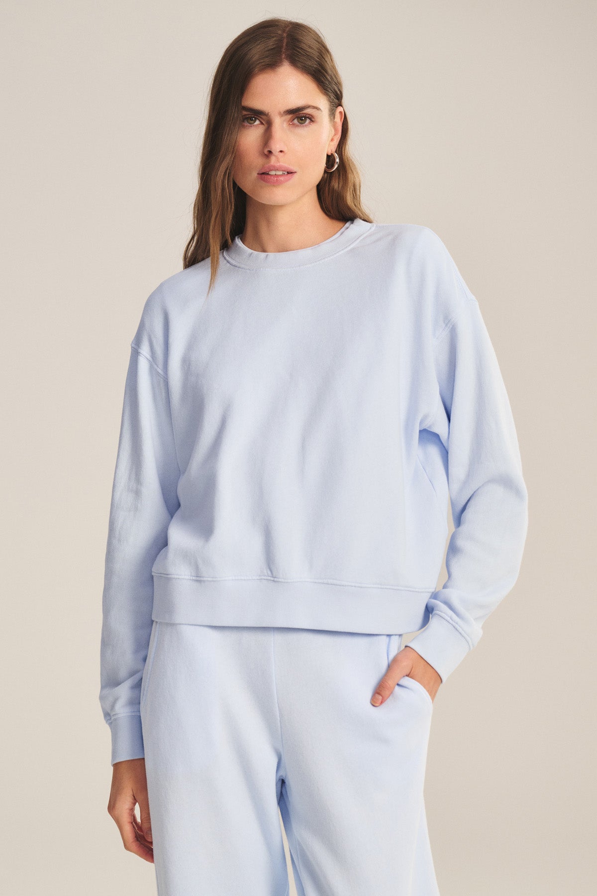A woman stands against a plain background, wearing the YOKO SWEATSHIRT by Velvet by Graham & Spencer in light blue soft cotton fleece with matching sweatpants, one hand in her pocket.-41044199768257