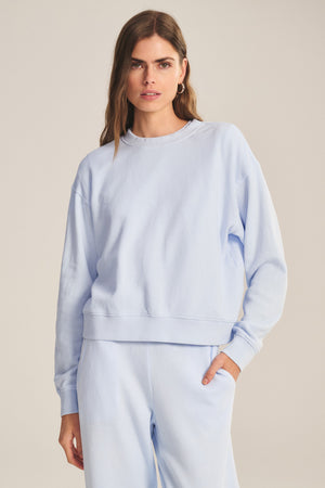 A woman stands against a plain background, wearing the YOKO SWEATSHIRT by Velvet by Graham & Spencer in light blue soft cotton fleece with matching sweatpants, one hand in her pocket.