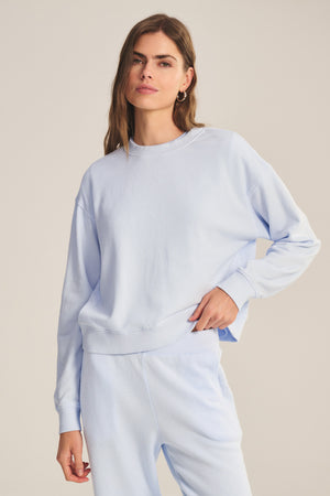 A woman models the YOKO SWEATSHIRT by Velvet by Graham & Spencer with matching light blue cotton fleece sweatpants, standing against a plain background and highlighting the relaxed fit.