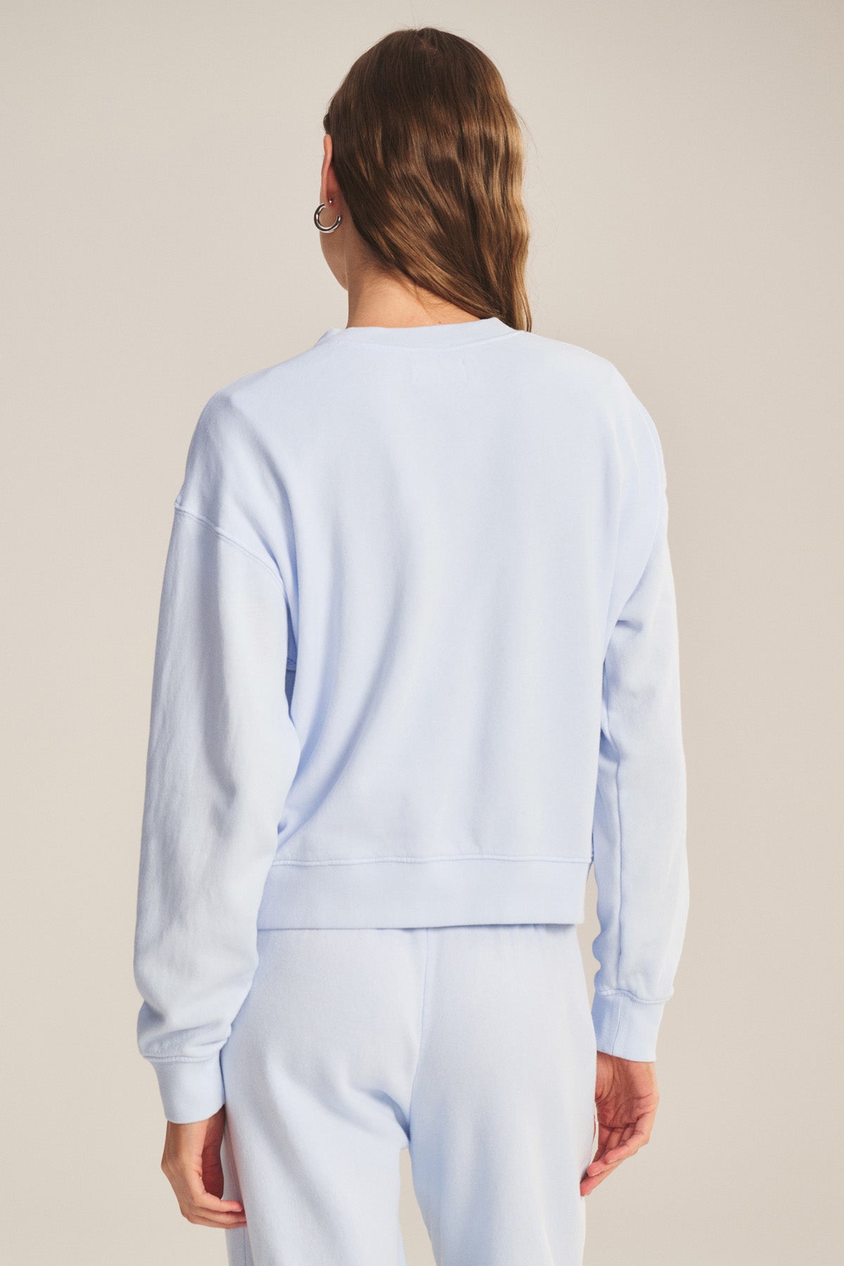 A person stands with their back to the camera in a light blue YOKO SWEATSHIRT by Velvet by Graham & Spencer, paired with matching sweatpants in soft cotton fleece, displaying a relaxed silhouette against a neutral backdrop.-41044199702721