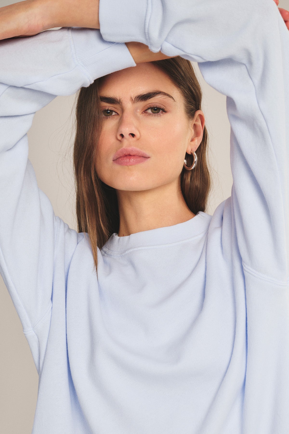 A woman wears the YOKO SWEATSHIRT by Velvet by Graham & Spencer, made from soft cotton fleece in light blue. With one arm raised, she faces the camera, her calm expression mirroring the sweatshirt's relaxed fit.-41044199669953