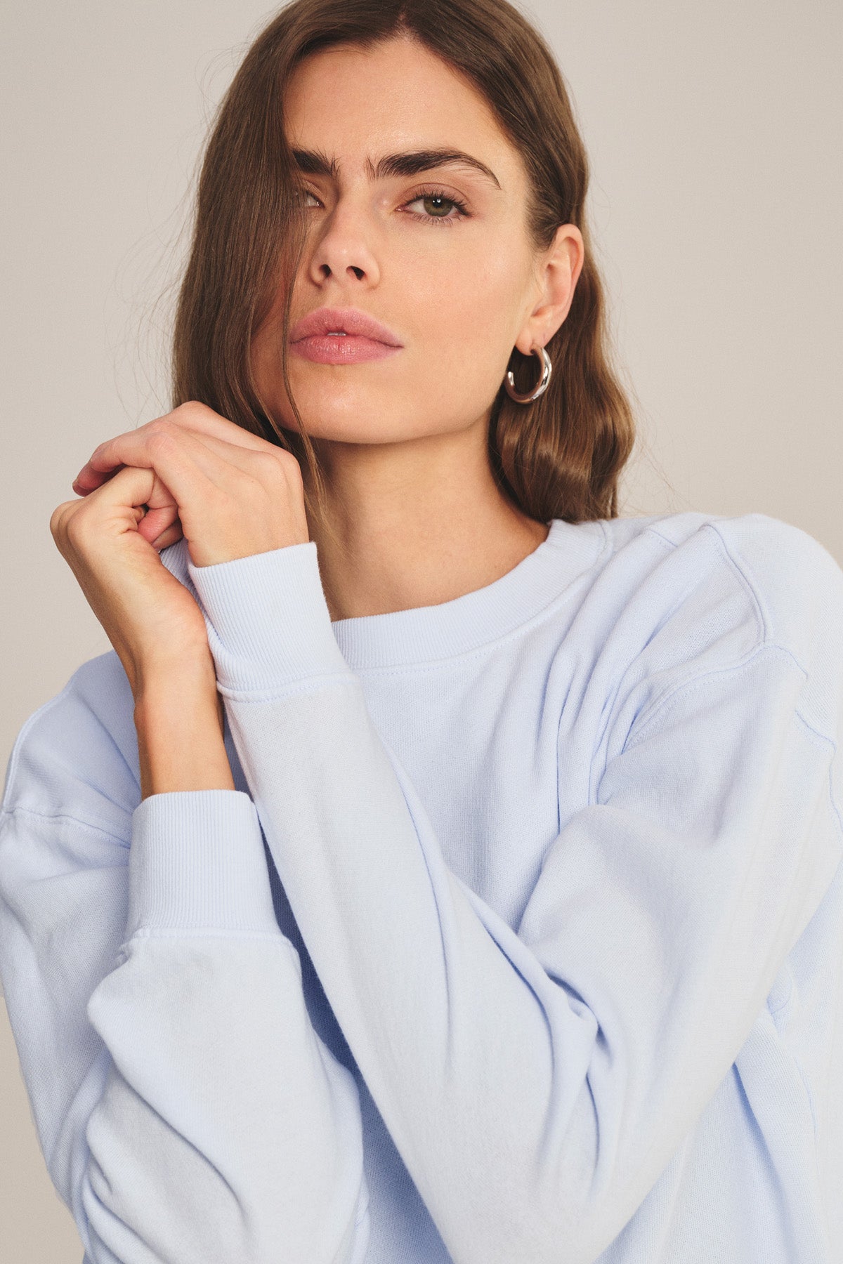 A woman with long brown hair wears the Velvet by Graham & Spencer YOKO SWEATSHIRT in light blue cotton fleece and silver hoop earrings, posing with her hands clasped near her face.-41044199604417