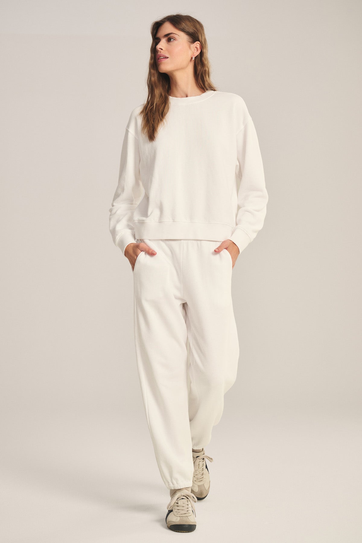 A person stands against a plain background, hands in pockets, wearing a relaxed fit white sweatshirt, ZARA SWEATPANT by Velvet by Graham & Spencer made from cotton fleece, and beige sneakers.