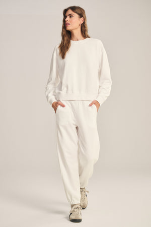 A person stands against a plain background, hands in pockets, wearing a relaxed fit white sweatshirt, ZARA SWEATPANT by Velvet by Graham & Spencer made from cotton fleece, and beige sneakers.
