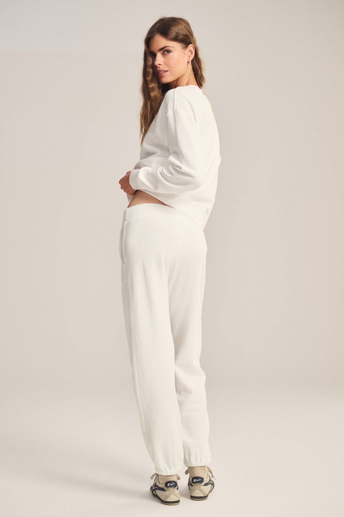 A woman stands facing slightly away, wearing a white cotton fleece sweatshirt, Velvet by Graham & Spencer ZARA SWEATPANT joggers, and sneakers against a plain background.-40431203483841