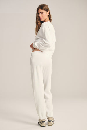A woman stands facing slightly away, wearing a white cotton fleece sweatshirt, Velvet by Graham & Spencer ZARA SWEATPANT joggers, and sneakers against a plain background.