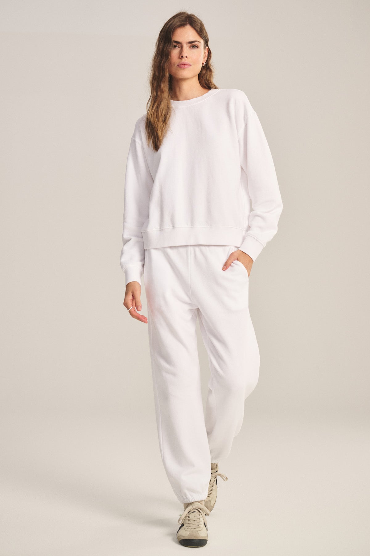 A person wearing the YOKO SWEATSHIRT by Velvet by Graham & Spencer and white sweatpants, with one hand in their pocket, walks forward on a plain background.