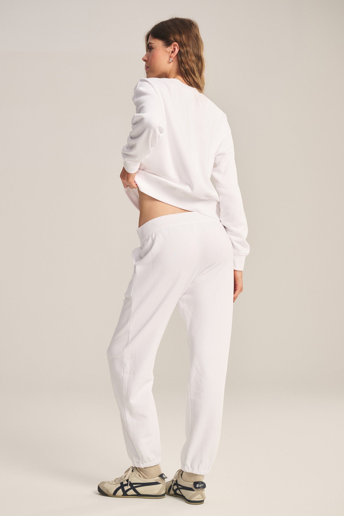 A woman faces away, wearing the YOKO SWEATSHIRT by Velvet by Graham & Spencer, paired with white sweatpants and white sneakers with black accents, set against a plain background.-40293969330369