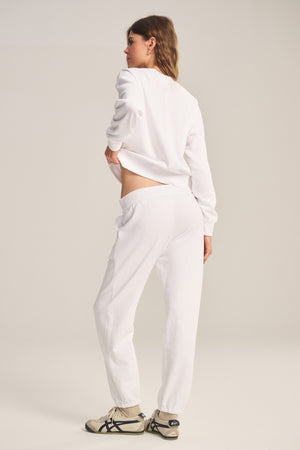 A woman faces away, wearing the YOKO SWEATSHIRT by Velvet by Graham & Spencer, paired with white sweatpants and white sneakers with black accents, set against a plain background.