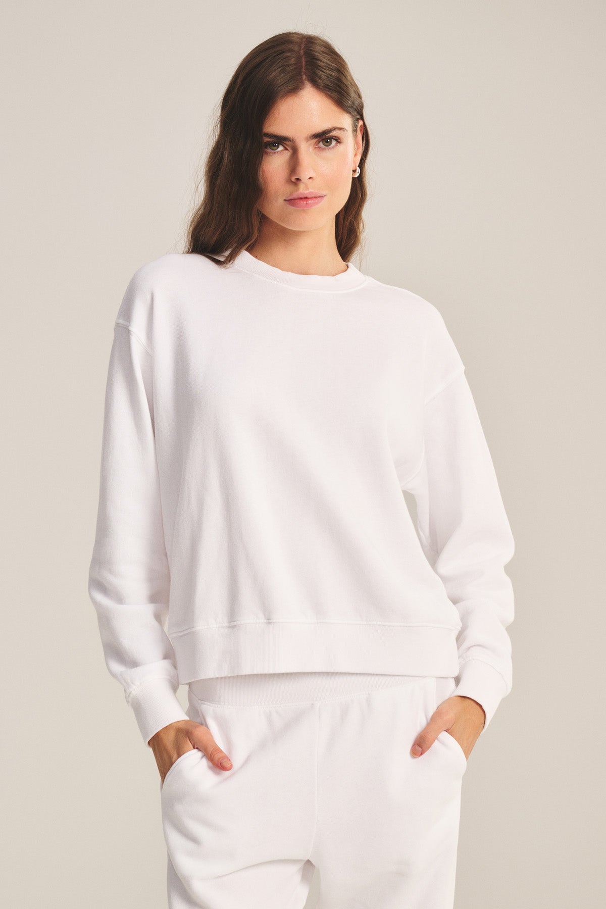 A woman stands facing forward, hands in pockets, wearing the Velvet by Graham & Spencer YOKO SWEATSHIRT with matching white pants against a neutral background.