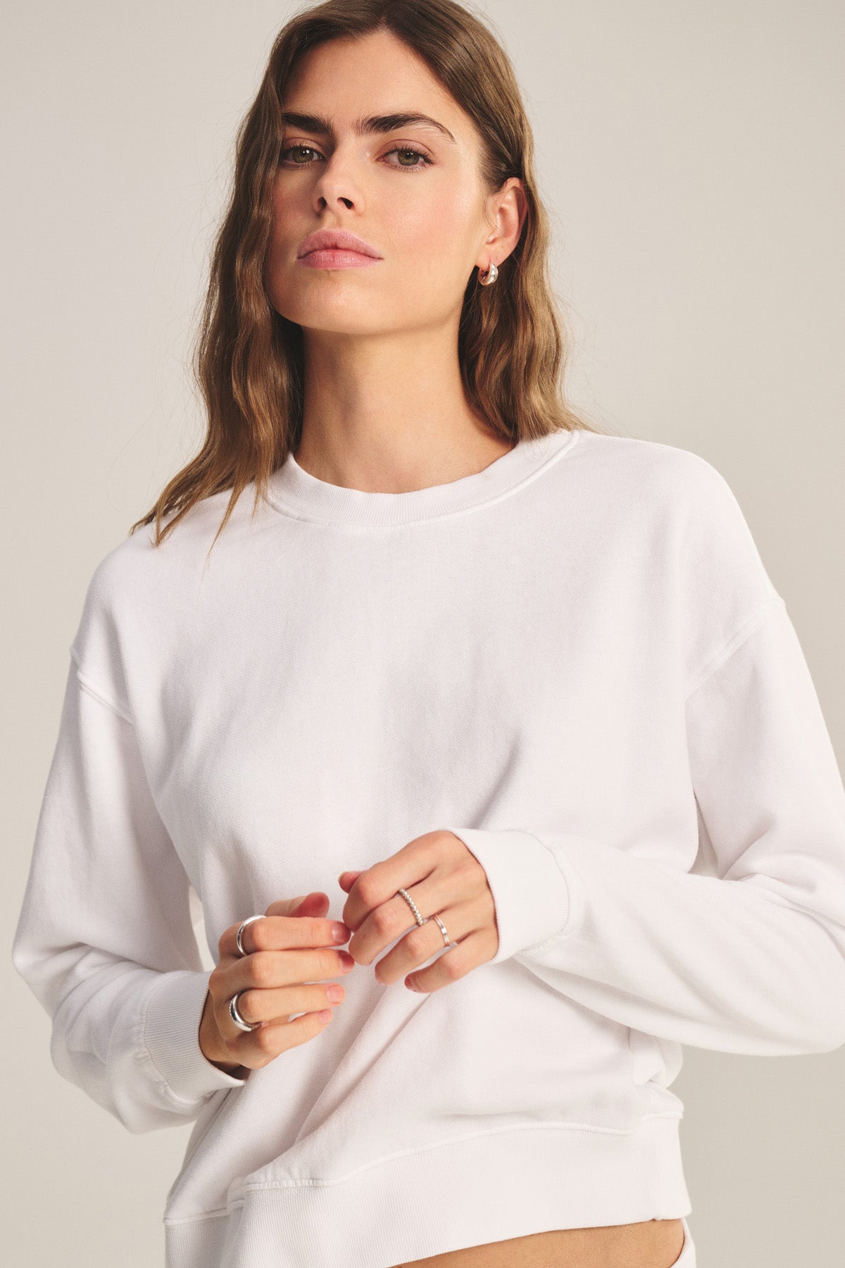 Woman with wavy brown hair wears the YOKO SWEATSHIRT by Velvet by Graham & Spencer in plain white, gazing at the camera with a neutral expression against a light background.