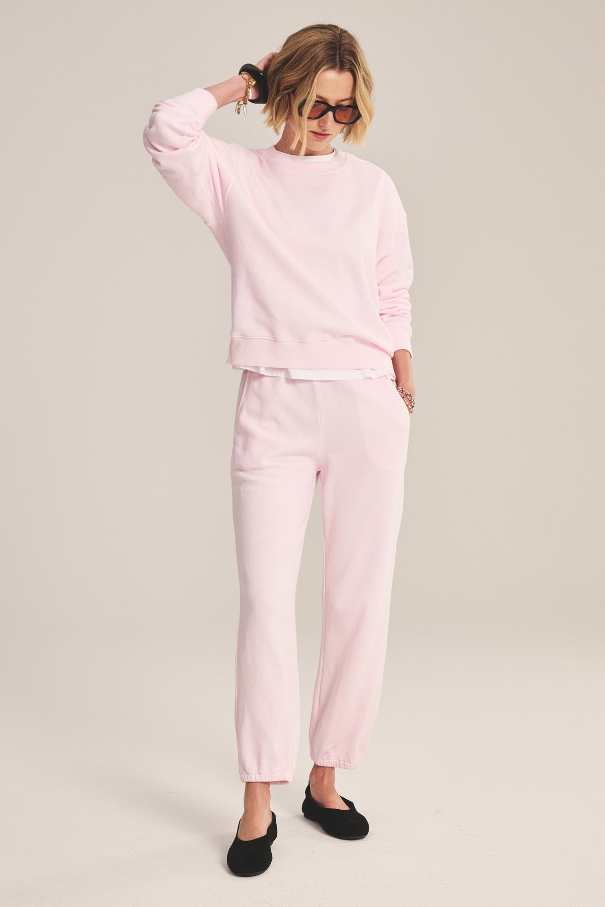 Wearing a relaxed silhouette, a person dons a light pink YOKO SWEATSHIRT by Velvet by Graham & Spencer with matching joggers, sunglasses, and black slip-on shoes, standing with one hand on the back of their head.