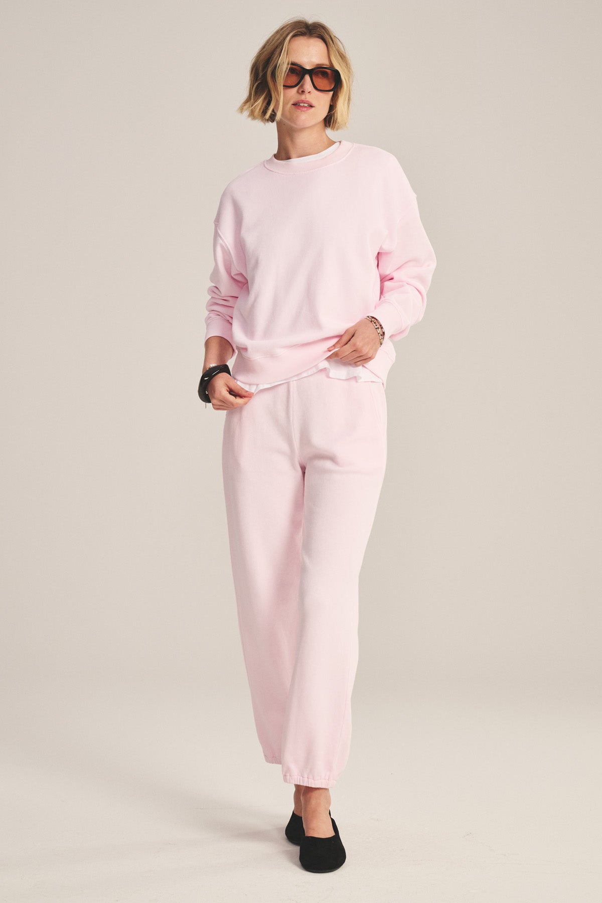 A person wears the Velvet by Graham & Spencer YOKO SWEATSHIRT in light pink with matching sweatpants, sunglasses, and black shoes. The cotton fleece offers extra comfort for an effortlessly cool, relaxed look against a plain background.-41044199473345