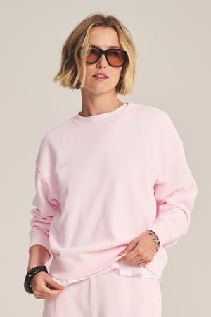 Person wearing a light pink cotton fleece YOKO SWEATSHIRT by Velvet by Graham & Spencer, matching light pink pants, and large brown sunglasses, with short blonde hair, standing against a plain background.