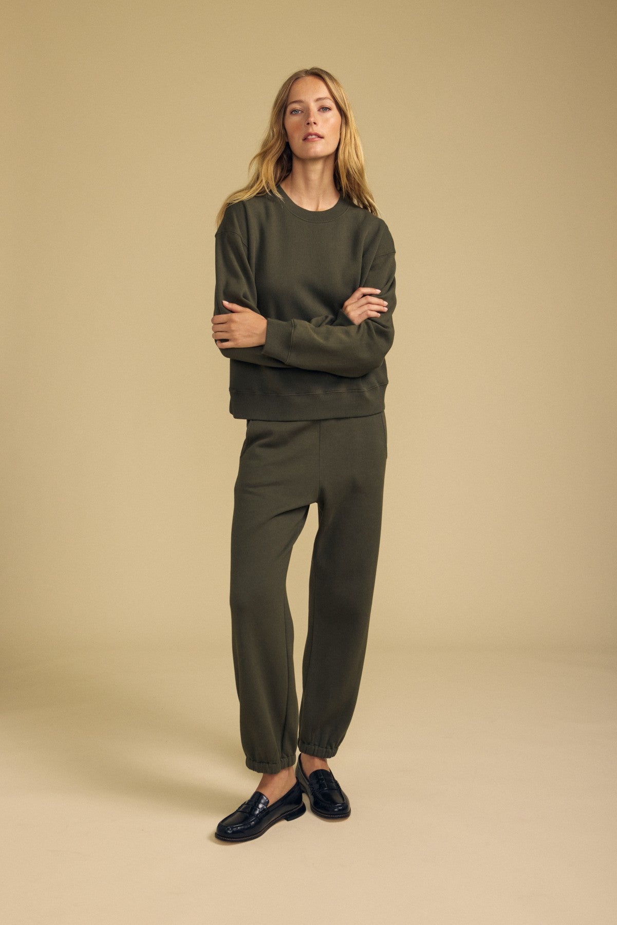 A woman stands in a neutral studio, wearing an olive green cotton fleece sweatshirt paired with the matching ZARA SWEATPANT jogger pants by Velvet by Graham & Spencer, styled with black loafers.