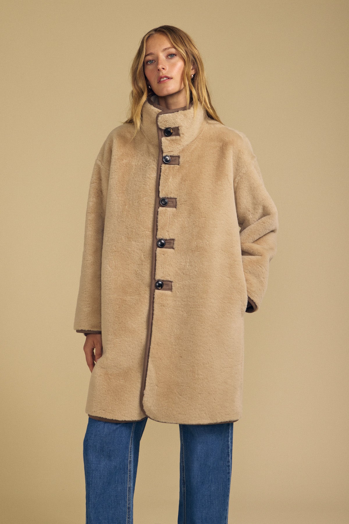 A person wearing the Velvet by Graham & Spencer YVES FAUX SUEDE FUR REVERSIBLE COAT—an oversized, buttoned beige coat with a high collar—paired with blue jeans stands against a plain beige background.-42063858270401