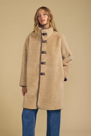 A person wearing the Velvet by Graham & Spencer YVES FAUX SUEDE FUR REVERSIBLE COAT—an oversized, buttoned beige coat with a high collar—paired with blue jeans stands against a plain beige background.