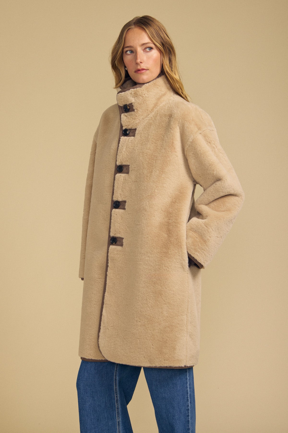 A woman models the YVES FAUX SUEDE FUR REVERSIBLE COAT by Velvet by Graham & Spencer—an elegant beige coat detailed with dark buttons, styled here with blue jeans against a plain beige background. This stylish piece features plush faux fur on one side and smooth faux suede on the other, offering versatile sophistication for any occasion.