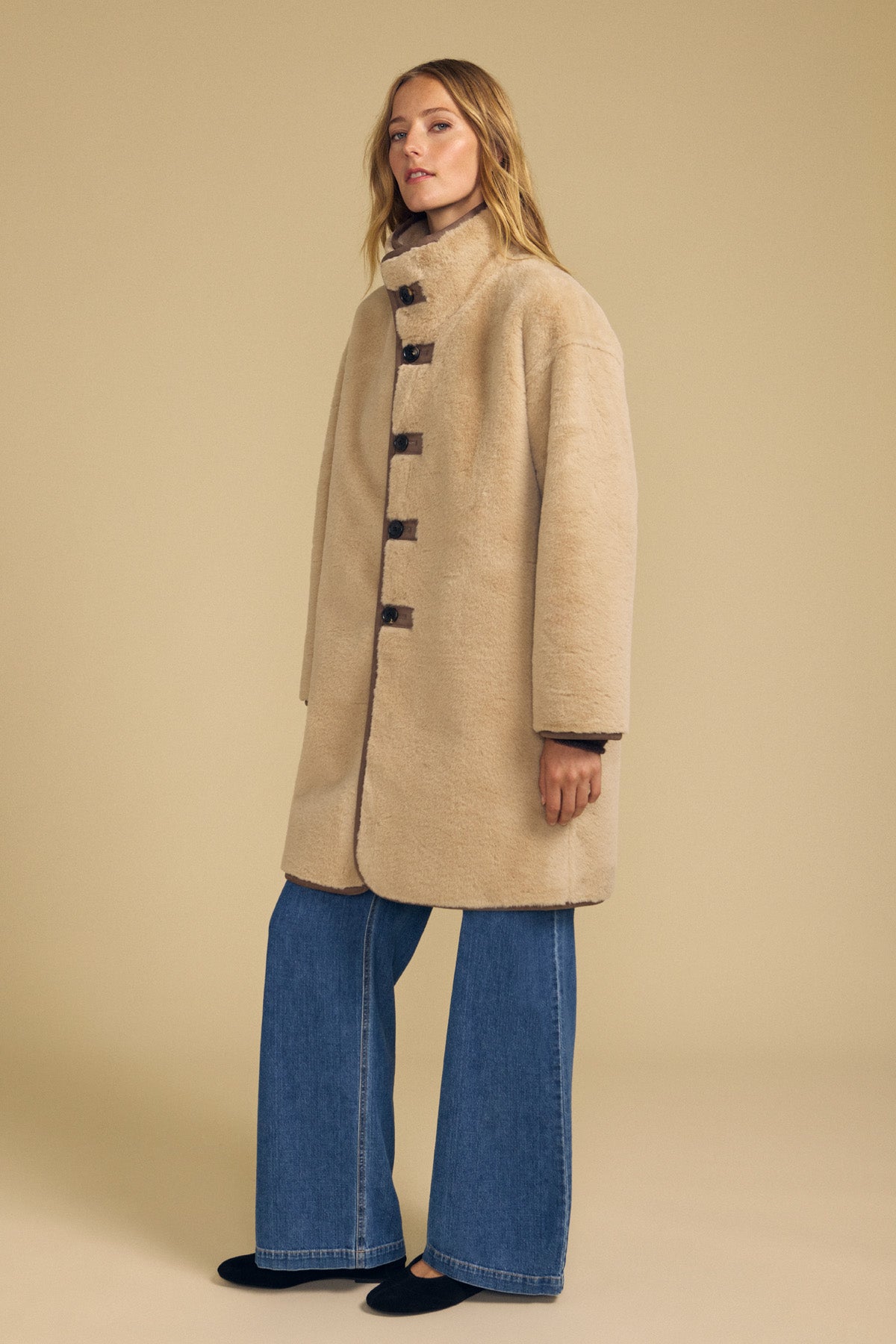 A woman models the Velvet by Graham & Spencer YVES Faux Suede Fur Reversible Coat—a beige, high-collar style with toggle closures crafted from luxurious faux suede—paired with wide-leg blue jeans and black shoes against a plain beige background.
