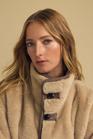 A woman with wavy blonde hair wears the YVES FAUX SUEDE FUR REVERSIBLE COAT by Velvet by Graham & Spencer, featuring large buttons, and looks at the camera against a plain beige background.