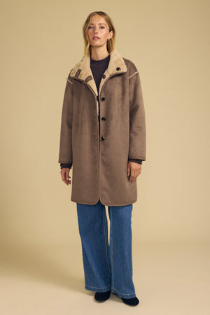 A woman stands against a beige background wearing the Velvet by Graham & Spencer YVES Faux Suede Fur Reversible Coat in brown, layered over a dark top with wide-leg blue jeans and black shoes.