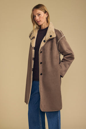 A person models the YVES FAUX SUEDE FUR REVERSIBLE COAT by Velvet by Graham & Spencer—featuring rich brown faux suede on one side and plush faux fur lining on the reverse—layered over a dark top and blue jeans, set against a plain beige background.