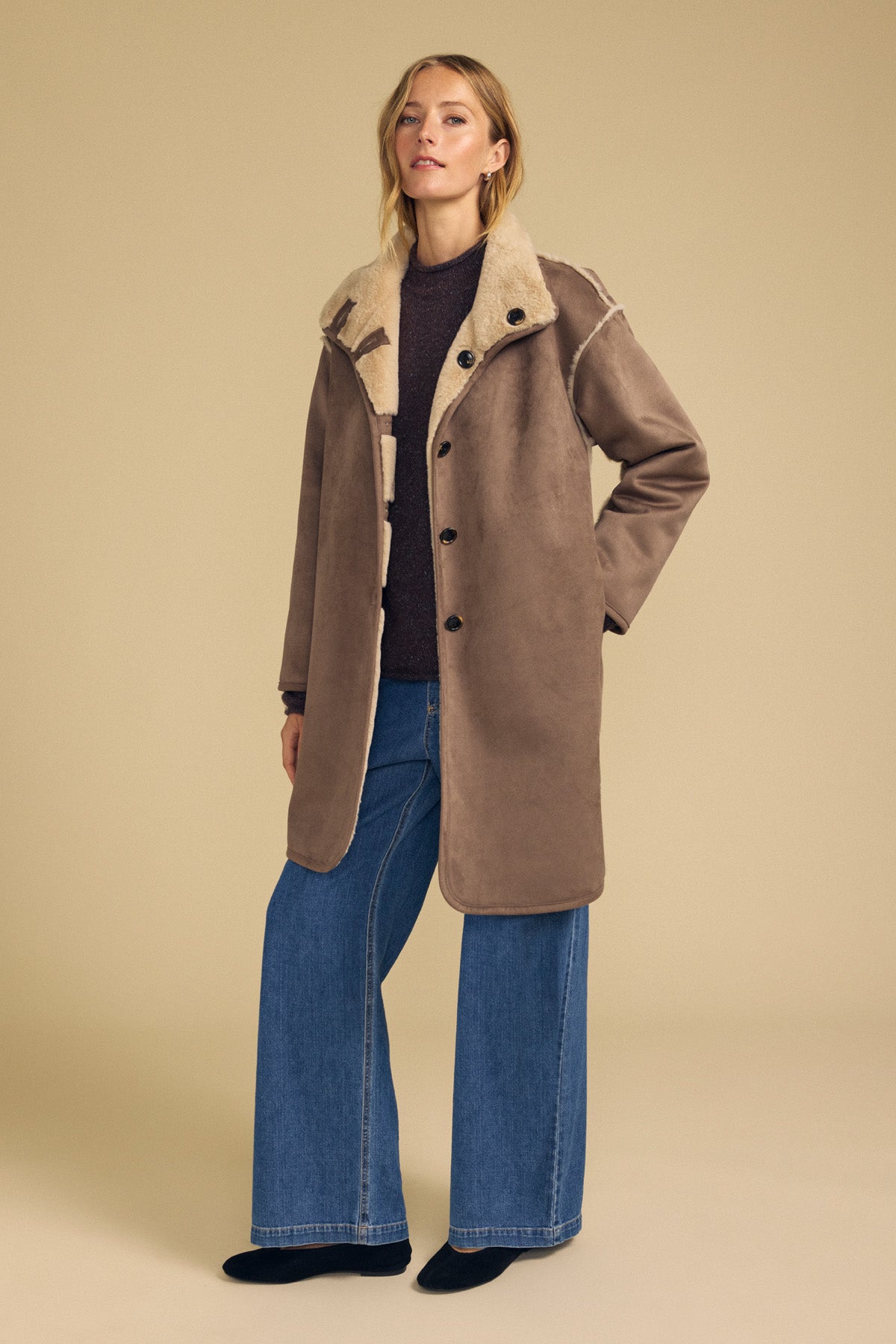 A woman models the YVES FAUX SUEDE FUR REVERSIBLE COAT by Velvet by Graham & Spencer, layered over a dark top and wide-leg blue jeans, set against a plain beige background.