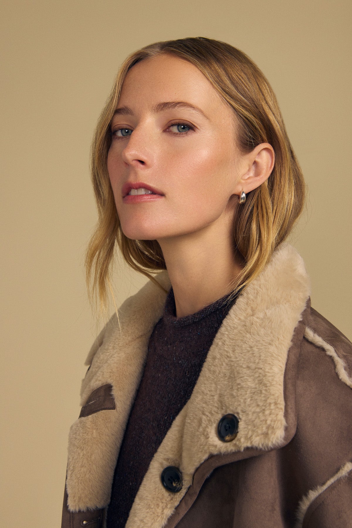 A woman with light brown hair wears the YVES FAUX SUEDE FUR REVERSIBLE COAT by Velvet by Graham & Spencer over a dark top, posing against a neutral beige background.