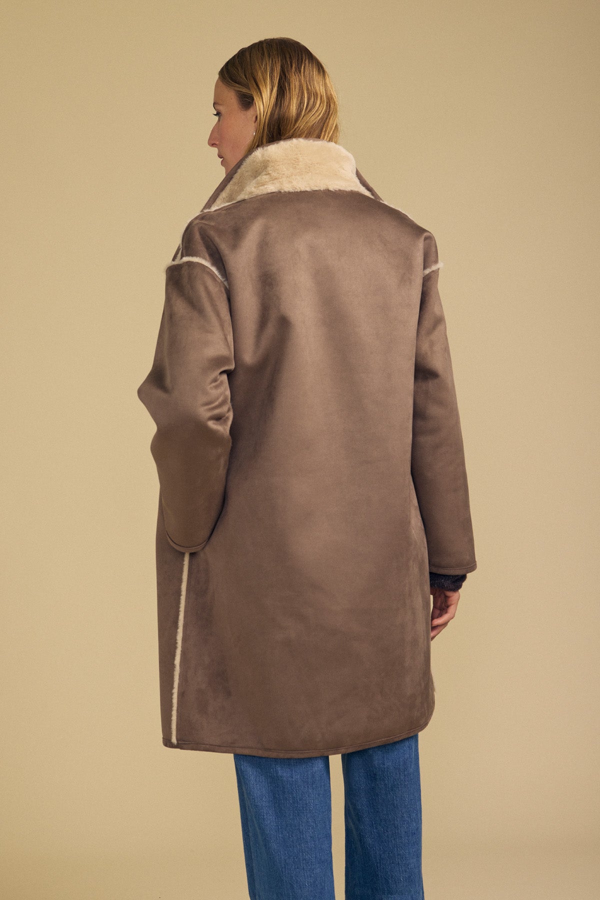 A person with long, light brown hair stands with their back to the camera against a beige background, wearing Velvet by Graham & Spencer’s YVES FAUX SUEDE FUR REVERSIBLE COAT featuring brown faux suede with light trim, paired with blue jeans.