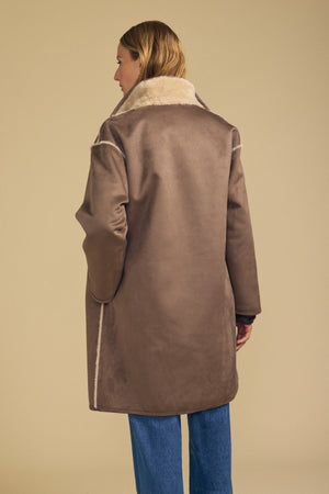 A person with long, light brown hair stands with their back to the camera against a beige background, wearing Velvet by Graham & Spencer’s YVES FAUX SUEDE FUR REVERSIBLE COAT featuring brown faux suede with light trim, paired with blue jeans.