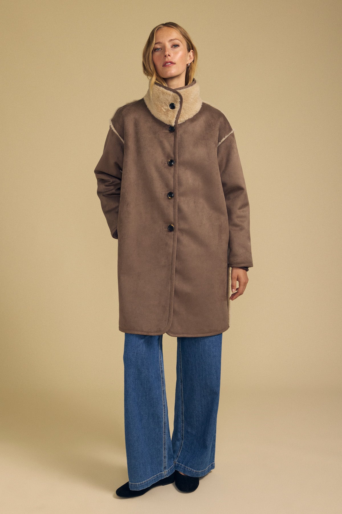 A person wearing the YVES FAUX SUEDE FUR REVERSIBLE COAT by Velvet by Graham & Spencer—a long brown faux suede coat with a high light-colored faux fur collar—paired with blue wide-leg jeans and black shoes stands against a plain beige background.-42063858237633