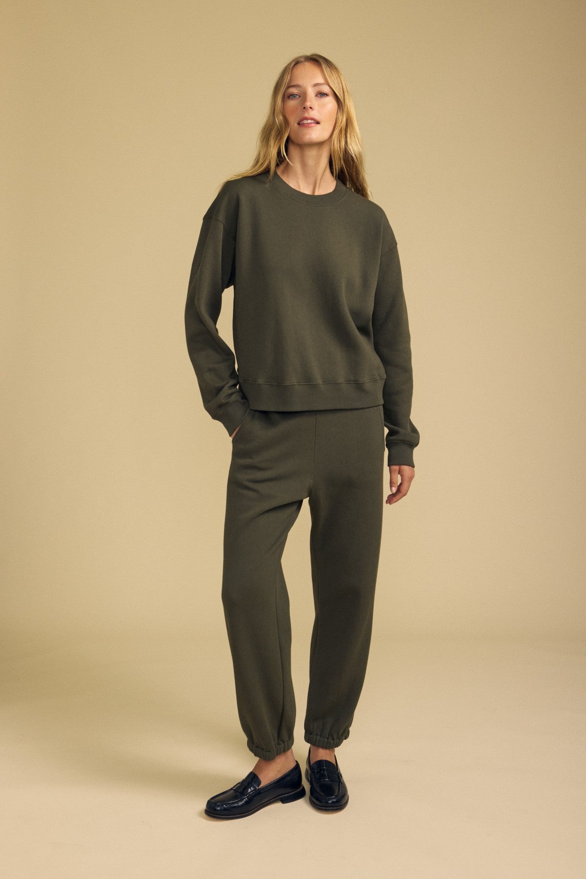 A woman stands on a neutral background wearing an olive green YOKO SWEATSHIRT by Velvet by Graham & Spencer and matching jogger pants, crafted from soft cotton fleece with a relaxed silhouette, paired with black loafers.