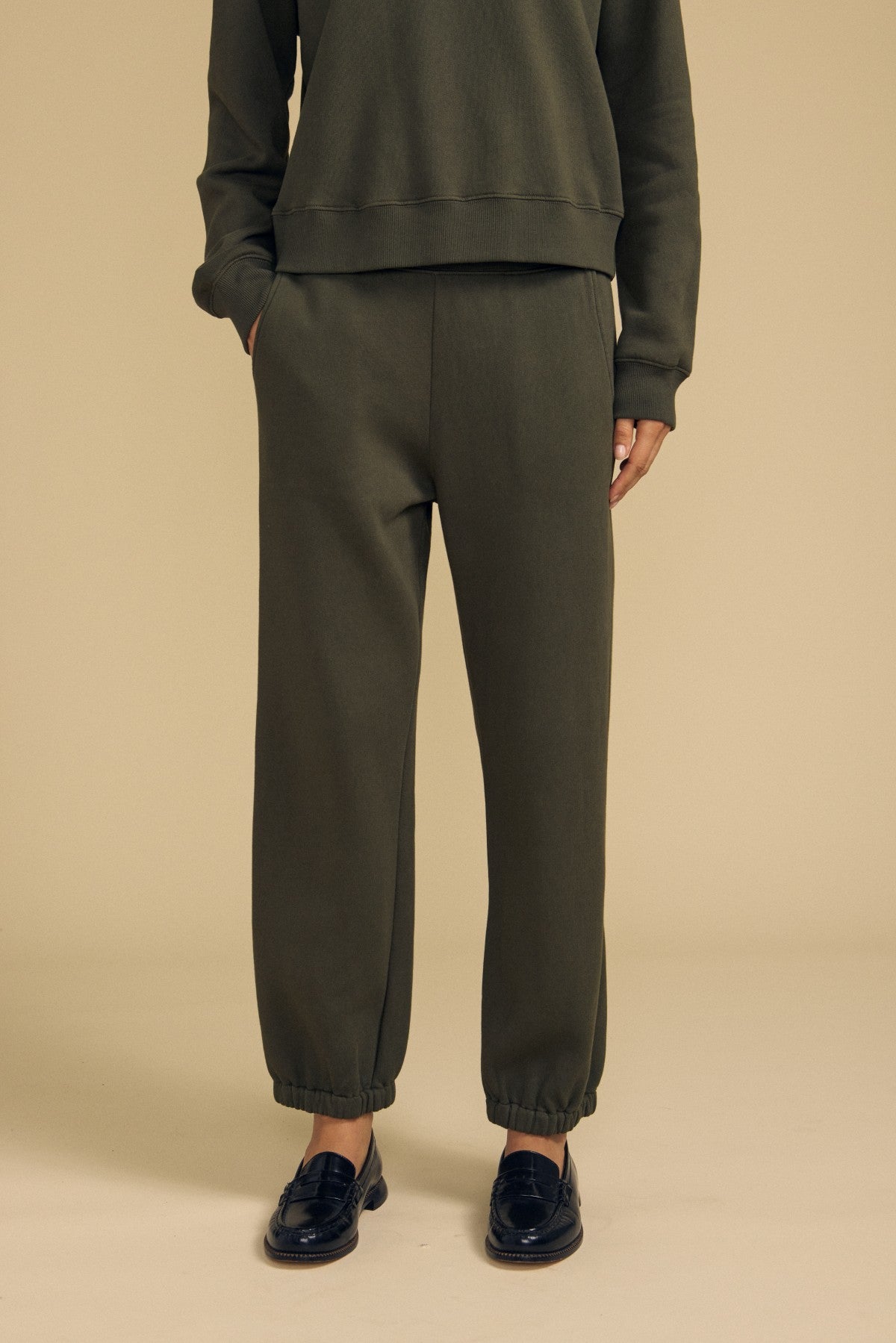 Wearing a dark green cotton fleece sweatshirt and Velvet by Graham & Spencer’s ZARA SWEATPANT jogger pants with a relaxed fit, paired with black loafers, the person stands against a plain beige background.